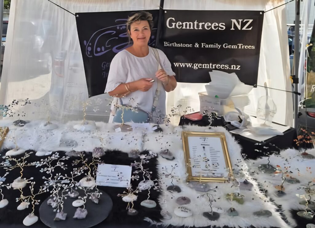 Woman standing at a Gemtrees NZ booth with a black banner reading 'Birthstone & Family Gem Trees' behind her.
