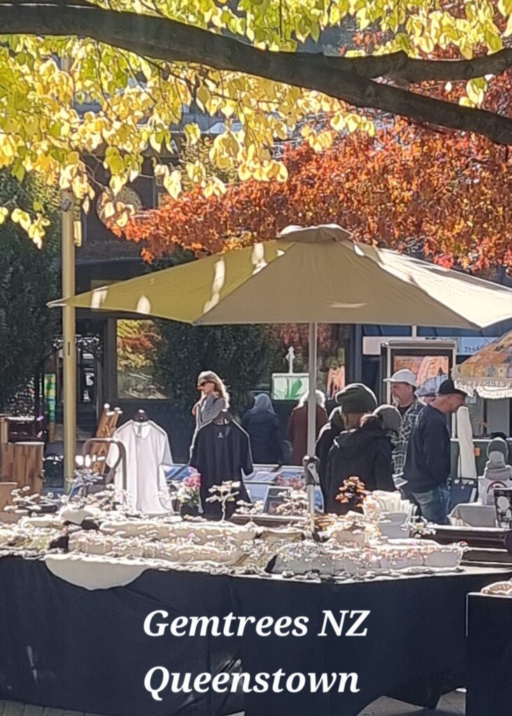 Outdoor market stall under yellow autumn leaves, displaying jewelry on tables with shoppers nearby.